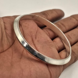 May include: A silver bangle bracelet with a smooth, polished surface. The bracelet is round and appears to be made of metal. The bangle is held in a hand, showcasing its design and shape. This piece of jewelry is suitable for everyday wear.