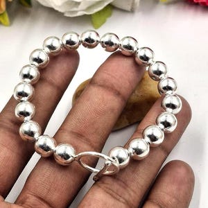 May include: A silver beaded bracelet with a toggle clasp. The bracelet features multiple round, shiny silver beads. The clasp is a simple, elegant design. The bracelet is shown on a person's hand.