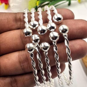 May include: A set of six silver-colored bangles. Each bangle features a twisted design and a small, round ball at the top. The bangles are displayed on a hand, with a blurred background of flowers.