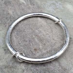 May include: A silver adjustable bangle bracelet. The bracelet is round with a hammered texture and a simple clasp. The bracelet has a small loop with three rings attached. The bracelet is on a grey surface.