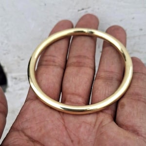 Pode incluir: Uma pulseira de argola dourada. A pulseira circular é segurada em uma mão, mostrando sua superfície lisa e polida. A pulseira é uma peça de joalheria simples e elegante.