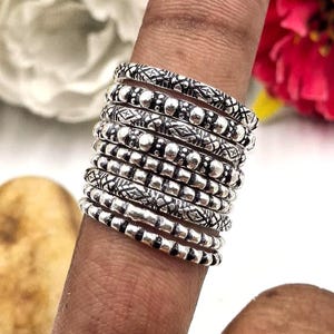 Puede incluir: Una pila de anillos de plata con intrincados diseños, incluyendo diseños de cuentas y grabados. Los anillos se llevan en un dedo, mostrando los detalles de cada banda. Los anillos son una mezcla de texturas y estilos.