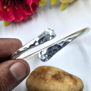 Puede incluir: Un brazalete plateado con textura martillada. El diseño abierto presenta dos terminales ensanchados. El brazalete se muestra sobre un fondo blanco con una flor roja y una piedra marrón.