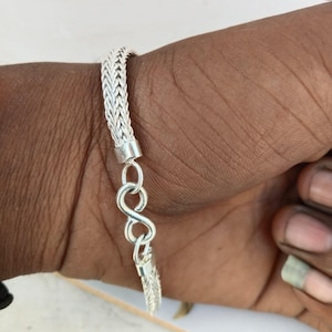 May include: A silver bracelet with a woven chain design. The bracelet has an infinity symbol clasp. The bracelet is shown on a person's wrist.