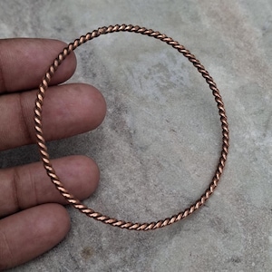 Op de afbeelding: Een koperkleurige, gedraaide metalen armband. De armband heeft een touwachtig ontwerp en is cirkelvormig. Het metaal heeft een warme, roodbruine tint. De armband wordt in een hand gehouden tegen een neutrale achtergrond.