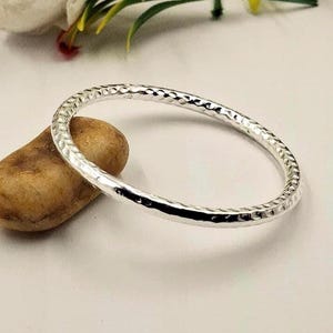 May include: A hammered silver bangle bracelet with a textured surface. The bracelet is circular and rests on a small, smooth, brown stone. The background is a soft white, with hints of green and yellow.