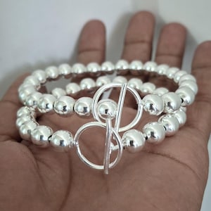 May include: Two silver beaded bracelets with a toggle clasp. The bracelets are made of round, shiny silver beads. The clasp is a silver circle with a bar that fits through it. The bracelets are shown on a person's hand.