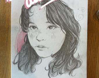 Op de afbeelding: Een potloodschetsportret van een persoon met donker haar en sproeten, met de naam "Millie" in roze schrift eronder. De woorden "little doodles" staan boven het portret in een cursief lettertype met een roze omlijning.