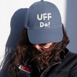 May include: A blue baseball cap with the text "UFF Da!" in white block letters. The hat is worn by a person, and the brim casts a shadow. The person is wearing a black jacket.