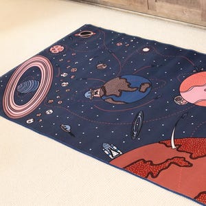 May include: A rectangular rug with a dark blue background featuring a colorful space-themed design. Planets, stars, and a rocket ship are depicted in shades of red, pink, brown, and white. The rug is placed on a light beige surface.