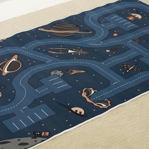 May include: A navy blue play mat with a space theme. The mat features a road map design with planets, rockets, and other space-related illustrations. The mat has a white border and is placed on a beige surface.