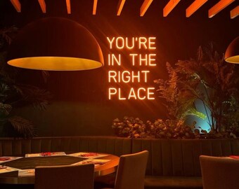 May include: Interior shot of a restaurant with a neon sign that reads "YOU'RE IN THE RIGHT PLACE." The sign is in bright orange against a dark background. The room has a warm, inviting atmosphere with round tables and chairs.