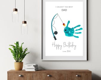 Könnte beinhalten: Gerahmter Druck mit dem Text "I Caught the Best Dad" und "Happy Birthday". Das Kunstwerk zeigt einen türkisfarbenen Handabdruck in Form eines Fisches mit einer Angelrute. Der Druck wird auf einem Holzschrank ausgestellt.