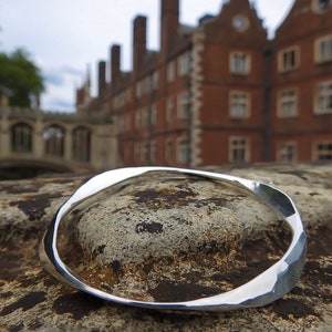 May include: A silver hammered bangle bracelet, lying on a rough, textured surface. The bracelet is thick and has a slightly irregular shape.