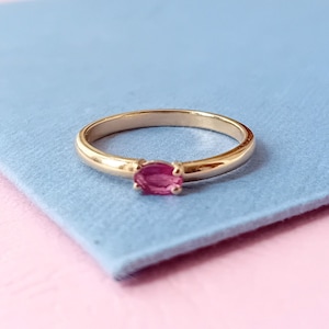 May include: A gold ring with a pink oval gemstone set in the centre.