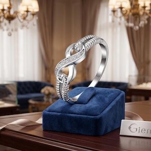 May include: A silver-toned ring featuring an infinity symbol design encrusted with small clear stones. The ring is displayed on a blue velvet stand next to a small silver plaque with the text Giemi engraved on the front surface of the metal plate.