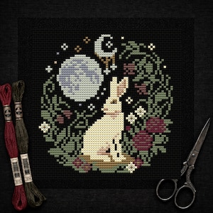 Könnte beinhalten: Ein Kreuzstichmuster mit einem weißen Hasen unter einem Vollmond und einer Mondsichel, umgeben von Blumen und Blättern. Das Design befindet sich auf schwarzem Hintergrund, mit Stickgarn und einer Schere im Rahmen.