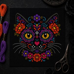 May include: A cross-stitch pattern on black fabric featuring a cat face decorated with vibrant floral motifs in orange, purple, pink, and blue. The cat has large yellow eyes, a pink heart-shaped nose, and white whiskers, surrounded by colorful flowers.