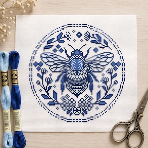 Könnte beinhalten: Ein quadratisches Stück weißer Aida-Stoff mit einem blauen Kreuzstichmuster einer Biene, das in einem kreisförmigen Blumenkranz zentriert ist. Zwei Stränge blaues Stickgarn und eine Metallschere liegen neben dem Stoff.