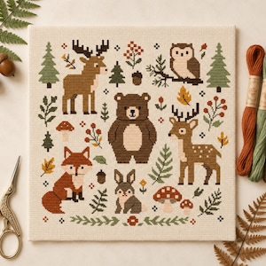 May include: A square cross-stitch embroidery on cream fabric featuring a woodland scene with a brown bear, a deer, a fox, a rabbit, an owl, and a moose. The design includes green pine trees, brown mushrooms, acorns, and orange leaves in a grid layout.