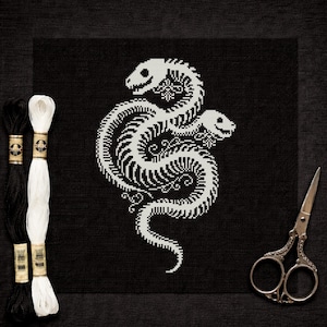 Könnte beinhalten: Ein schwarzes Kreuzstichdesign mit einem weißen Schlangenskelett mit zwei Köpfen. Das Bild enthält schwarzes und weißes Stickgarn und eine dekorative Schere. Das Schlangendesign ist auf einem schwarzen Stoff zentriert.