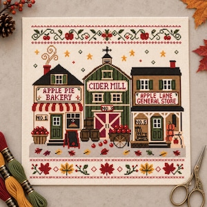 May include: A cross-stitch pattern on cream fabric featuring three buildings: Apple Pie Bakery, Cider Mill, and Apple Lane General Store. The design includes red apples, barrels, a cart, and autumn leaves, framed by decorative borders of apples and leaves.