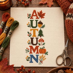 May include: A cross-stitch pattern on cream-coloured Aida fabric featuring the word AUTUMN spelled vertically. Each letter is accompanied by seasonal motifs including a maple leaf, acorn, pine tree, mushroom, pumpkin, and wheat stalks in autumn colours.