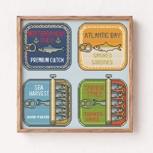 Könnte beinhalten: Ein gerahmtes Kreuzstich-Kunstwerk mit vier Sardinenbüchsen-Designs. Jede Dose ist mit Texten wie "Mediterranean Coast" und "Atlantic Bay" beschriftet. Die Dosen sind in Blau-, Orange- und Grüntönen gehalten und zeigen Fischillustrationen.
