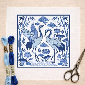 May include: A cross-stitch design of two blue cranes, bamboo, clouds, and lotus flowers. The design is framed in a square on white fabric. Two spools of blue embroidery thread and a pair of scissors are also visible. The design uses shades of blue.