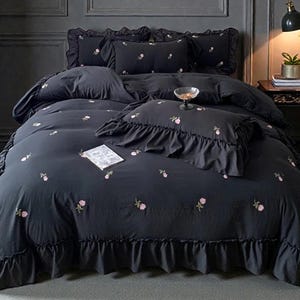 Puede incluir: Juego de cama gris oscuro con detalles florales rosas bordados. El juego incluye una funda nórdica, almohadas y un borde con volantes. Un pequeño cuenco decorativo y un libro están sobre la cama. La habitación tiene paredes grises oscuras.