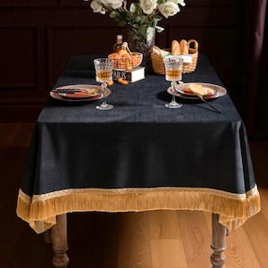 May include: A dark blue rectangular tablecloth with a gold fringe trim, set on a wooden table. The table is set with plates, glasses, and a basket of bread. A vase of white roses and a small basket of oranges are also on the table.