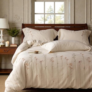 Puede incluir: Un juego de cama color crema con un estampado floral. El juego incluye una funda nórdica y fundas de almohada, todas adornadas con delicados tallos florales verticales en una paleta de colores suaves y apagados. La cama está contra un cabecero de madera y una ventana.