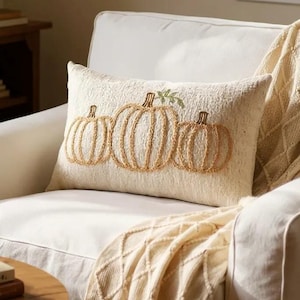 Op de afbeelding: Een rechthoekig crèmekleurig kussen met drie geborduurde pompoenen in tinten beige en bruin. Het kussen ligt op een witte fauteuil, met een crèmekleurige deken over de arm. Een kopje koffie staat op een kleine houten tafel.