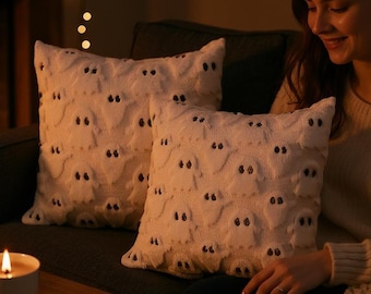 May include: Two cream-colored throw pillows with ghost face designs rest on a dark gray sofa. A lit candle in a white mug, a plate of cookies, and a book sit on a wooden table. A fireplace with a warm glow is in the background.