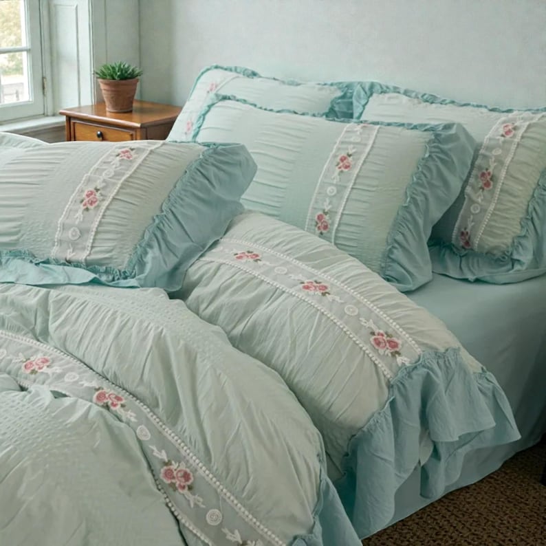 May include: A light blue bed set with ruffled edges and floral accents. The set includes pillows and a comforter, all featuring a delicate white lace trim with pink floral embroidery. The bedding is arranged on a bed in a room with a window.