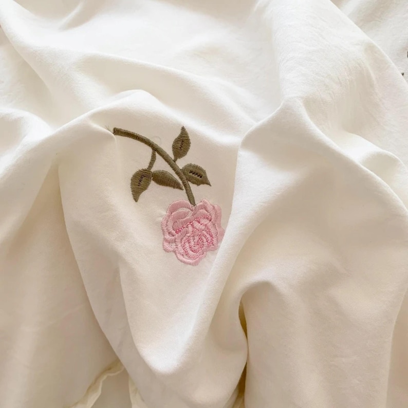 May include: Cream-colored fabric with a delicate embroidered pink rose and green leaves. The fabric is softly draped, creating gentle folds and shadows. The floral embroidery adds a touch of elegance and detail to the textile.