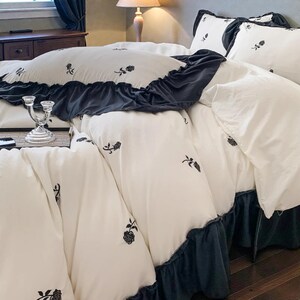 May include: White bedding set with black floral embroidery. The set includes a duvet cover with a dark gray ruffle trim, matching pillowcases, and a bed skirt. The bedding is arranged on a bed in a bedroom setting.