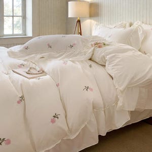 May include: White bedding set with embroidered pink floral accents. The set includes a duvet cover, pillowcases, and a bed skirt. A book and a pearl necklace rest on the bed. The room has a neutral color palette with a lamp and window in the background.