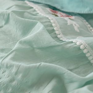 May include: Close-up of a light green fabric with a textured appearance. The fabric has a delicate embroidered section with floral designs in pink and white, bordered by a white pom-pom trim. The overall aesthetic is soft and airy.