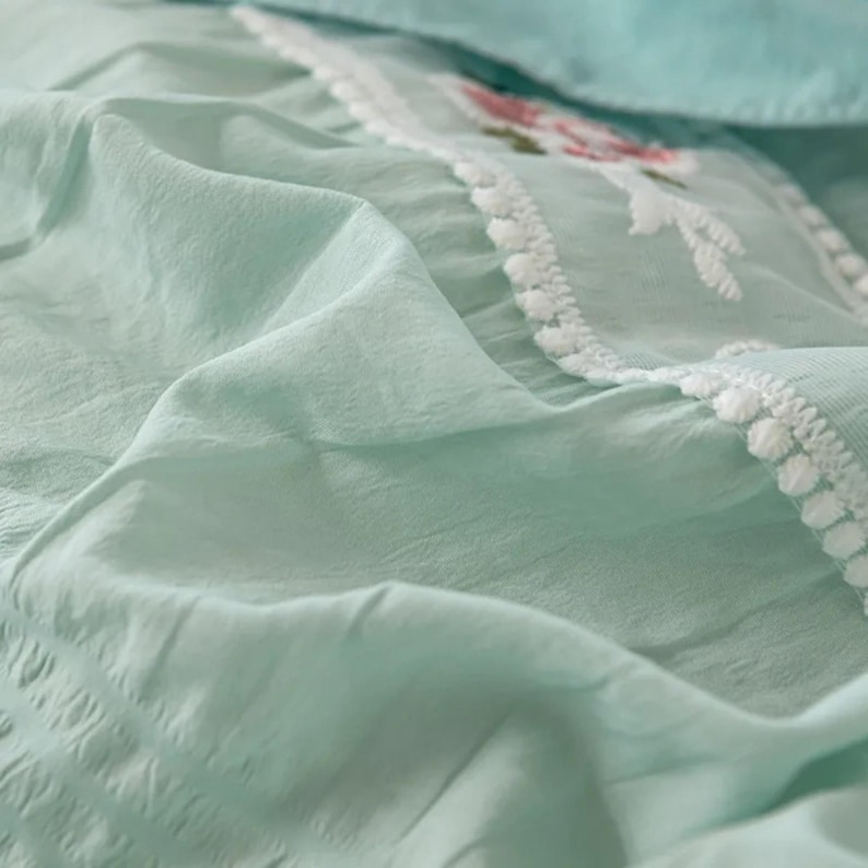 May include: Close-up of a light green fabric with a textured appearance. The fabric has a delicate embroidered section with floral designs in pink and white, bordered by a white pom-pom trim. The overall aesthetic is soft and airy.