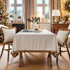 以下が含まれることがあります： クリーム色のフリンジ付きテーブルクロスのあるダイニングルームのシーン。クリーム色のクッションとフリンジエッジが付いた2つの椅子が見えます。クリスマスツリーと装飾品が、お祝いの雰囲気を醸し出しています。テーブルの長さは約150cmです。