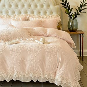 Puede incluir: Un lujoso juego de cama rosa con intrincados patrones bordados y ribetes de encaje. El juego incluye un edredón, almohadas y una falda de cama, todo en un estilo suave y romántico. La cabecera es un diseño capitoné en color crema.