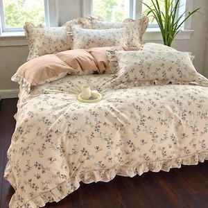 Puede incluir: Un juego de cama con estampado floral, con una funda nórdica y fundas de almohada de color crema adornadas con pequeñas flores en tonos azules, marrones y verdes. El juego incluye bordes con volantes y varias almohadas con estampados lisos y de lunares.