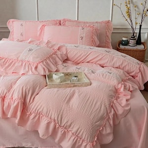 May include: A pink bedding set with ruffled edges and embroidered details. The set includes a comforter, pillowcases, and a bed skirt. A decorative tray with a white object and a small box sits on the bed. The bedding has a romantic, vintage aesthetic.