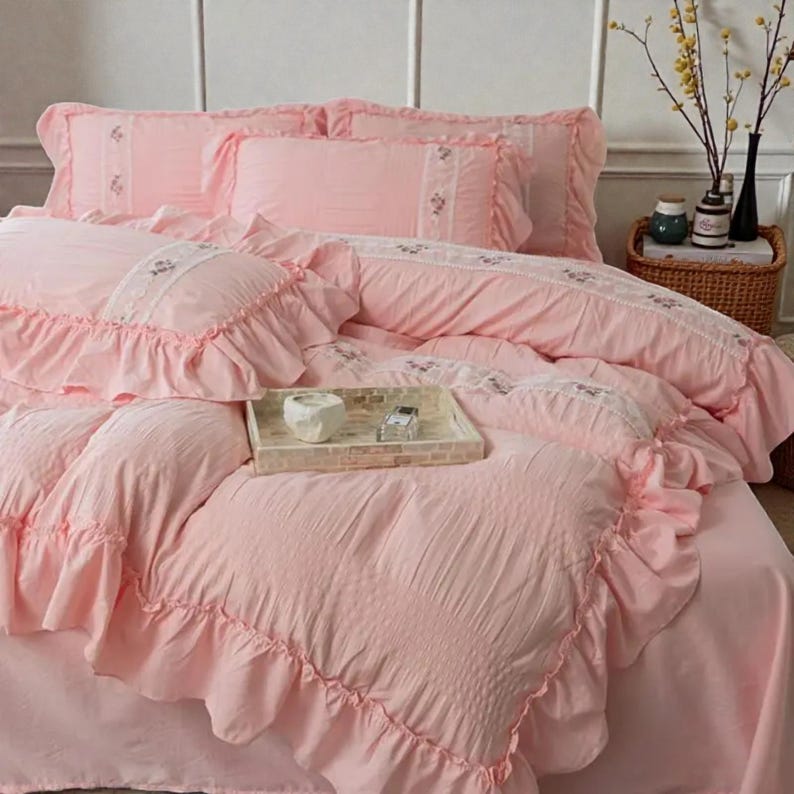 May include: A pink bedding set with ruffled edges and embroidered details. The set includes a comforter, pillowcases, and a bed skirt. A decorative tray with a white object and a small box sits on the bed. The bedding has a romantic, vintage aesthetic.