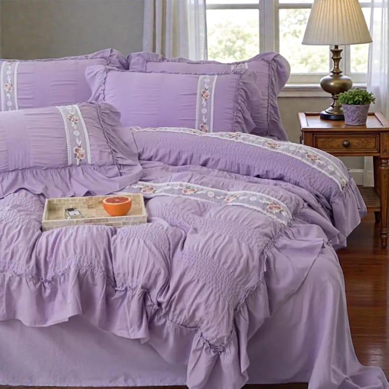 May include: A lavender-colored bedding set with ruffled edges and floral accents. The set includes a comforter, pillowcases, and a bed skirt. A wooden tray with a grapefruit sits on the bed. A lamp and side table are in the background.