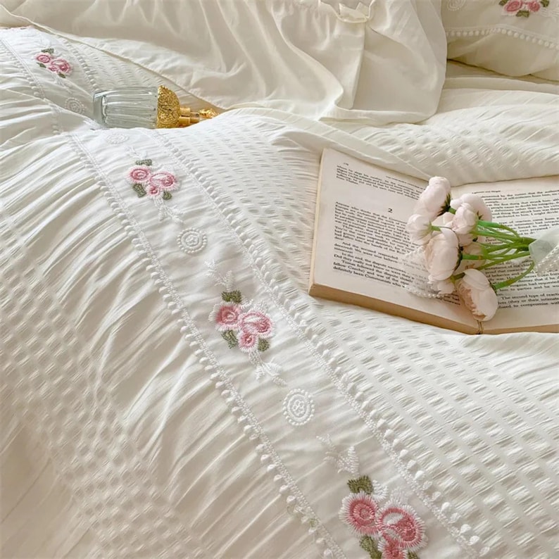 May include: Cream-colored bedding set with embroidered pink roses and green leaves. The set includes a duvet cover and pillowcases. A glass bottle and a book with a bouquet of pink flowers are also present.