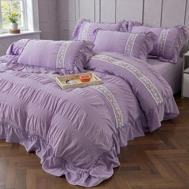 May include: A lavender bed set with ruffled edges and embroidered floral accents. The set includes a comforter, pillows, and a bed skirt. A wooden tray with a grapefruit sits on the bed. The bedding is made of a soft, textured fabric.