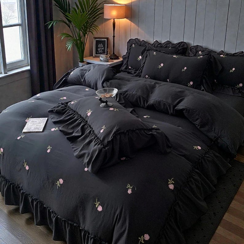 May include: A black bed set with a floral pattern. The set includes a comforter, pillowcases, and decorative pillows. The comforter and pillowcases have a ruffled edge. The floral pattern is a delicate pink color.