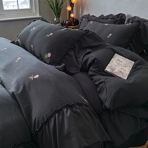 May include: Dark gray bedding set with embroidered pink roses. The set includes a comforter, pillows, and a long bolster pillow. The comforter and pillowcases have ruffled edges. A small book with a floral design rests on the comforter.